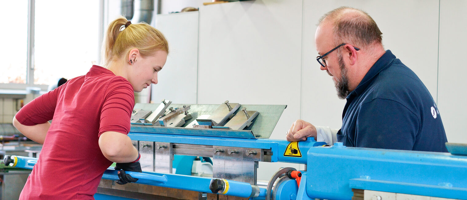 Spenglerin bei der Ausbildung Auszubildene Spenglering biegt an einer Maschine ein großes Blech. Ausbilder überwacht die Szene.