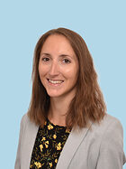 Bild von einer Frau mit langen braunen Haaren, grauem Jacket und schwaz, gelb geblümter Bluse vor hellblauem Hintergrund.